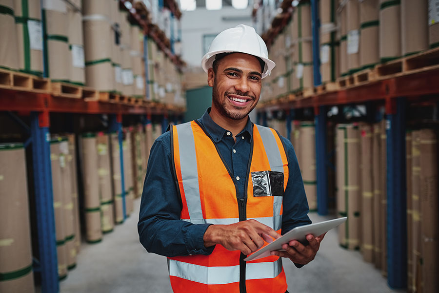 Warehousing and Logistics Insurance - Portrait of Smiling Warehouse Manager Using a Digital Tablet in a Warehouse While Standing between Shelves and Looking at the Camera