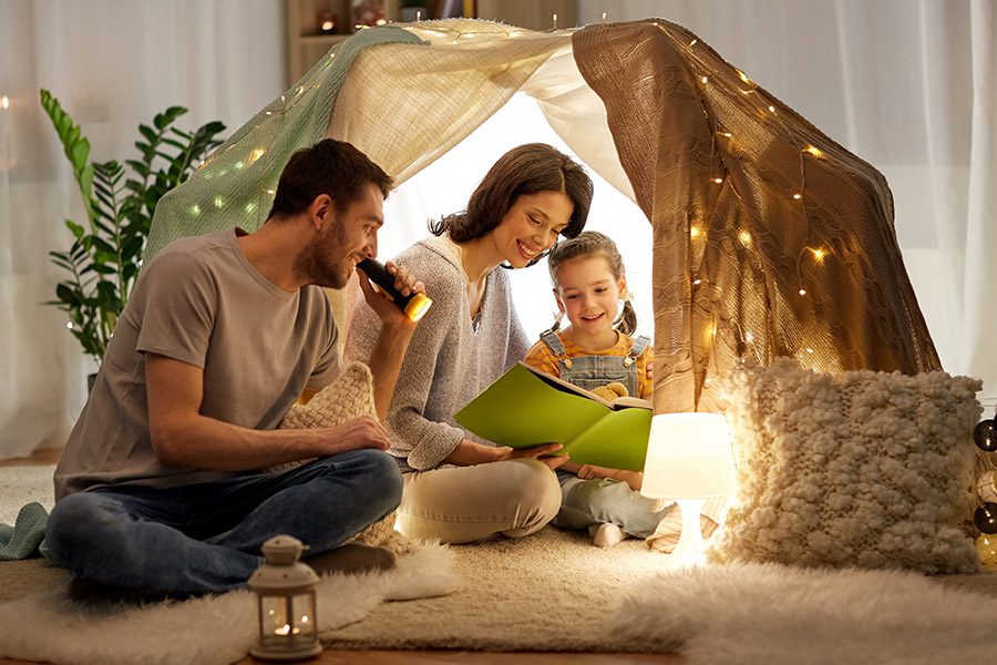 Personal Insurance - A Happy Family is Sitting in a Tent Covered in Lights With Their Daughter Reading a Storybook at Home