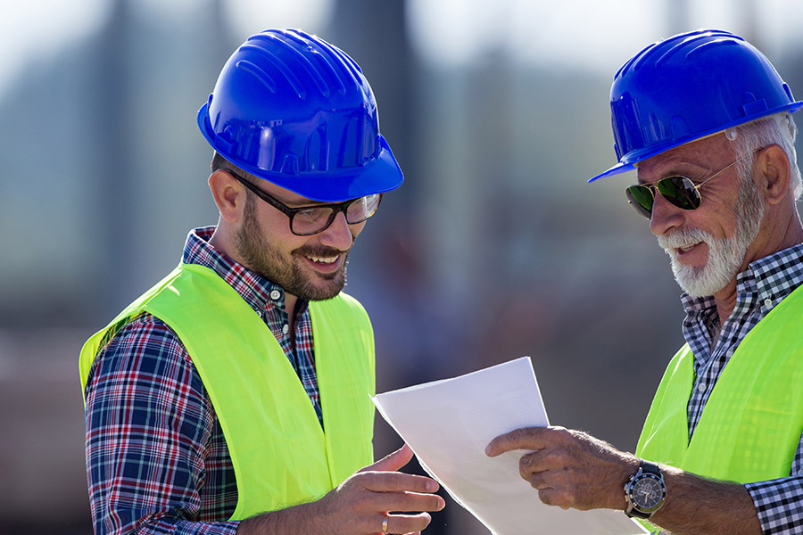 Specialized Business Insurance - Two Engineers Wearing Blue Hard Hats and Yellow Vests are at a Site Discussing Construction Plans on a Sunny Day